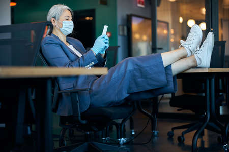 Elegant woman in medical mask using smartphone at workの写真素材