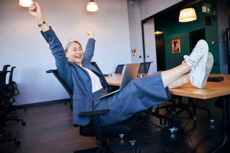 Cheerful woman using laptop and stretching at workの写真素材