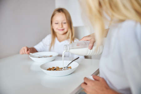 Adult woman pouring milk for their breakfast while she is sitting with little girl in the kitchen homeの写真素材