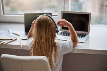 Cute small miss in casual clothes looking at the two screen her laptops while sitting at the white tableの写真素材
