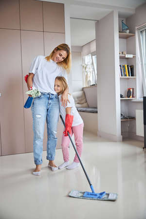 Happy cheerful small girl with woman in casual clothes standing with spray and floor mop in living roomの写真素材
