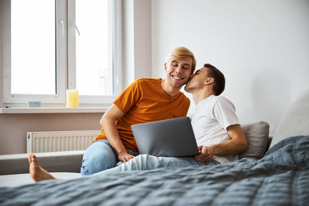 Happy gay couple cuddling, kissing and using laptop at homeの写真素材