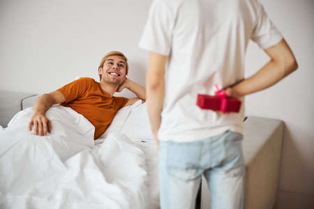 Cheerful young man lying in bed and looking at boyfriend with presentの写真素材