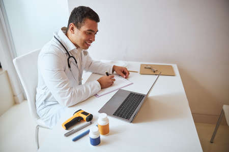 Young male general practitioner writing something on clipboard while sitting at the work desk in room indoorsの写真素材