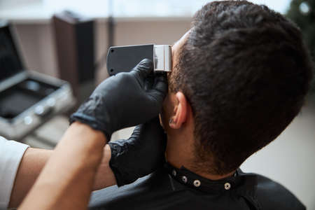 Hands of barber in medicine gloves holding modern machine and making undercut on man visitorの写真素材