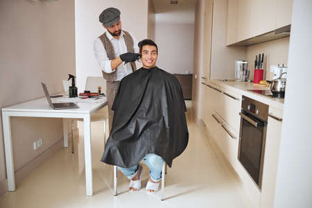 Stylish hair stylist in classic suit and cap making hair cut with special instrument in room indoors