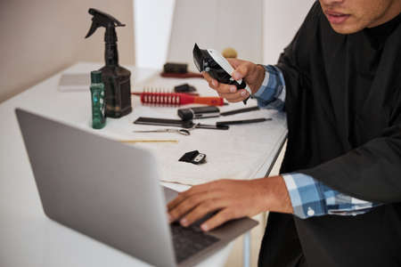 Focused man with different hair tools on the desk looking to the screen of laptop in home apartments