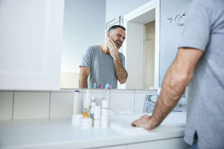 Unhappy young man looking in the mirror in bathroomの写真素材