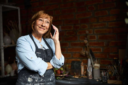 Joyful senior woman talking on cellphone in pottery workshop