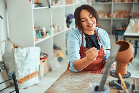 Cheerful young woman painting pottery in workshop