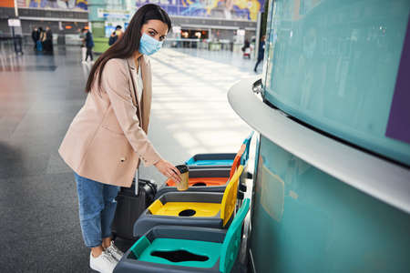 Nice woman in medical mask throwing coffee cup in trash can at airport