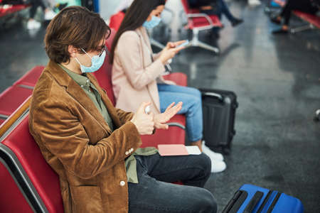 Stylish man in medical mask using hand sanitizer at airportの写真素材