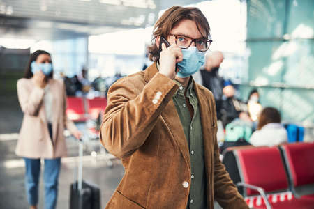 Young man in medical mask having phone conversation at airportの写真素材
