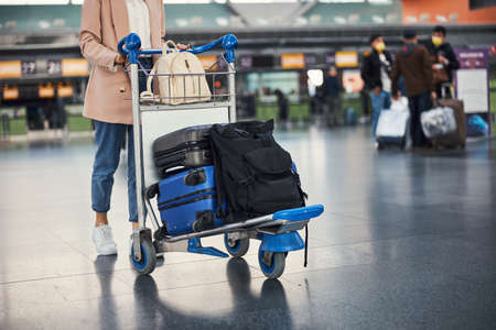 Stylish young woman pushing luggage trolley at airportの写真素材