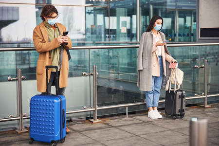 Young man and woman in medical masks using smartphones outdoorsの写真素材