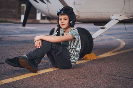 Adorable boy in aviation hat sitting near aircraft at aerodrome
