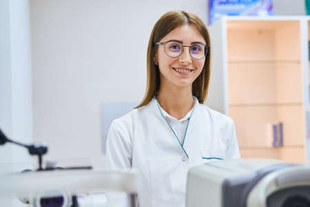 Cheerful female doctor standing by autorefractor in clinic