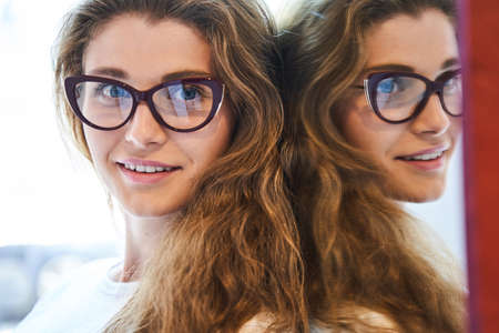 Beautiful young woman in glasses standing by mirrorの写真素材