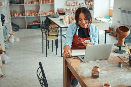 Joyful woman using laptop and smartphone in pottery workshop