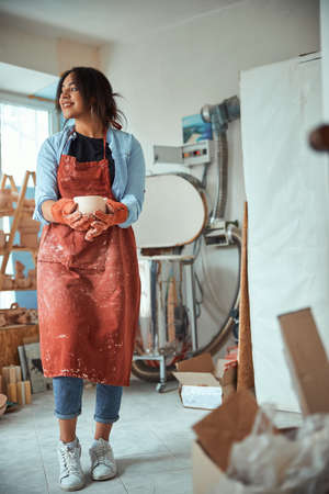 Charming female potter in apron holding handmade clay bowlの写真素材