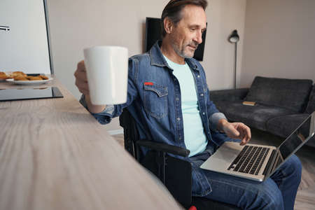 Smiling man on wheelchair with laptop drinking coffee