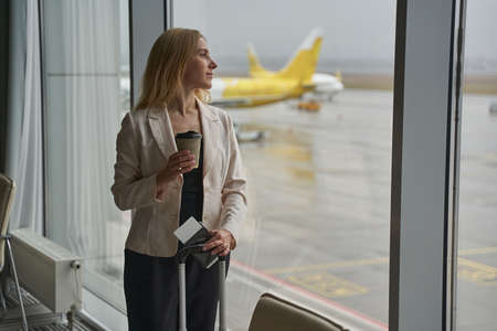 Woman drinking coffee while standing with luggage in airport terminalの写真素材