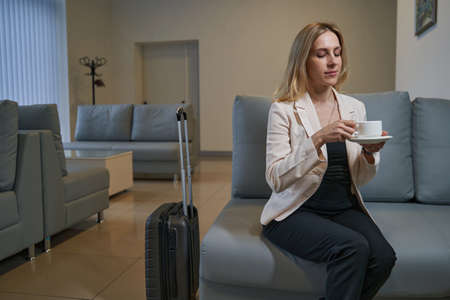 Woman sitting in hotel lobby with suitcase and coffeeの写真素材