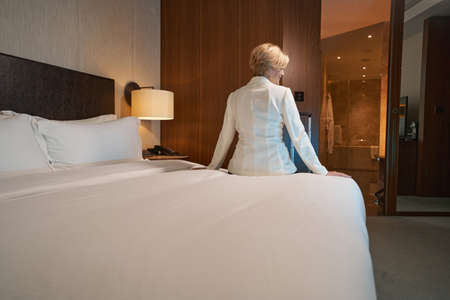 Rear-view photo of a blonde lady in white blazer sitting on hotel bed