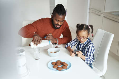 African American man making breakfast for his daughter while she playing on the smartphoneの写真素材