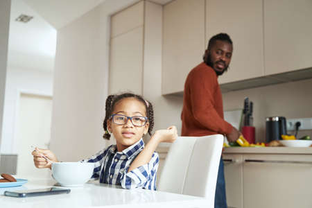 Joyful girl eating in the kitchen in the company of her African American fatherの写真素材