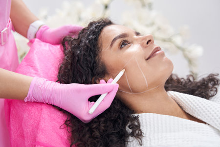 Beautician giving markings on cheek for beauty procedure for young woman
