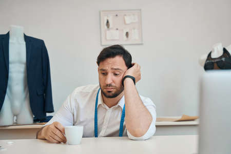 Man couturier sits at table in atelier holding a cup