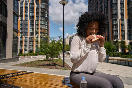 Multiracial female eating a sandwich while sitting on a bench