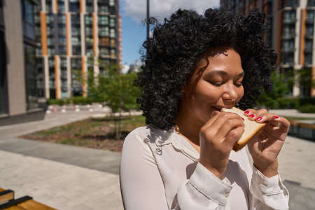 Multiracial female eats a sandwich on the background of tall housesの写真素材