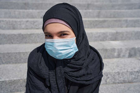 Woman in a hijab sits on the stairs wearing a maskの写真素材