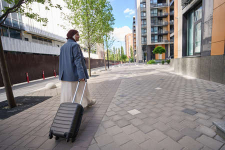 Woman in a hijab walks down the street with a suitcase on wheelsの写真素材