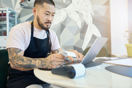 Calm cafe worker sorting out paperwork at table with laptopの写真素材