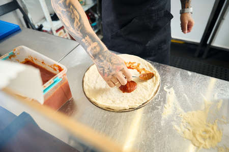 Professional pizza chef covering dough circle with red sauceの写真素材