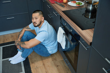 Thoughtful asian man sitting in the kitchen on the floorの写真素材