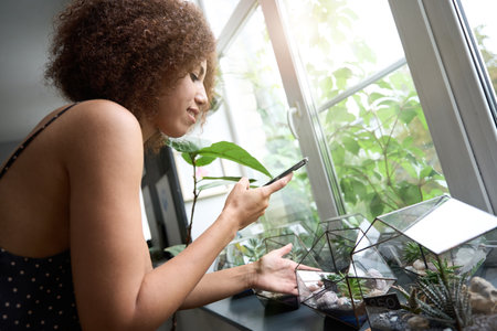 Joyous curly lady photogrpahing plats at her windowsillの写真素材