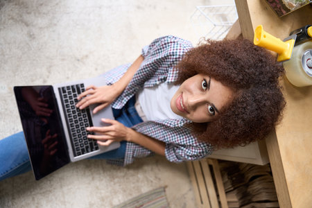 Cheerful curly woman using laptop in workshopの写真素材