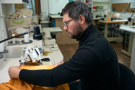 Male tailor works at overlocker, his workplace is well lit