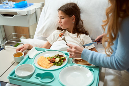 Mom helps her daughter in a hospital bed with a mealの写真素材