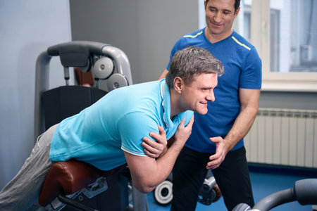 Mature man exercising on hyperextension bench supported by fitness instructor