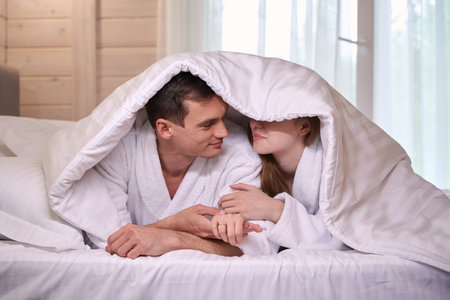 Happy couple in bathrobes with blanket covering heads on bedの写真素材