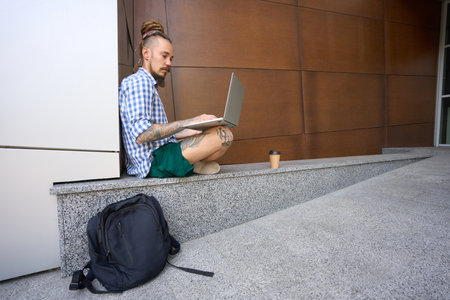 Informal guy works on laptop on granite curb near modern buildingの写真素材
