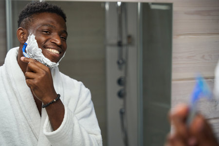African American guy shaves in front of mirror in bathroomの写真素材