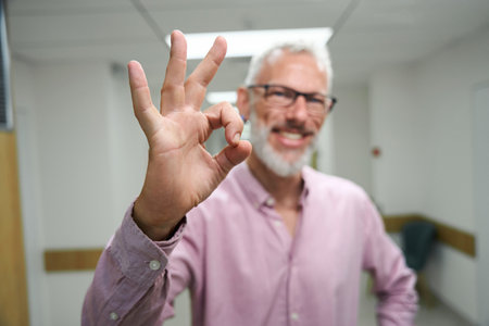 Bearded gray-haired man in a pink shirt shows ok