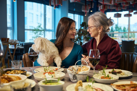 Young and aged women holding hands and talking sitting in restaurant with dog
