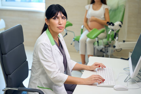 Gynecologist looking at camera while typing on computer and examining womanの写真素材